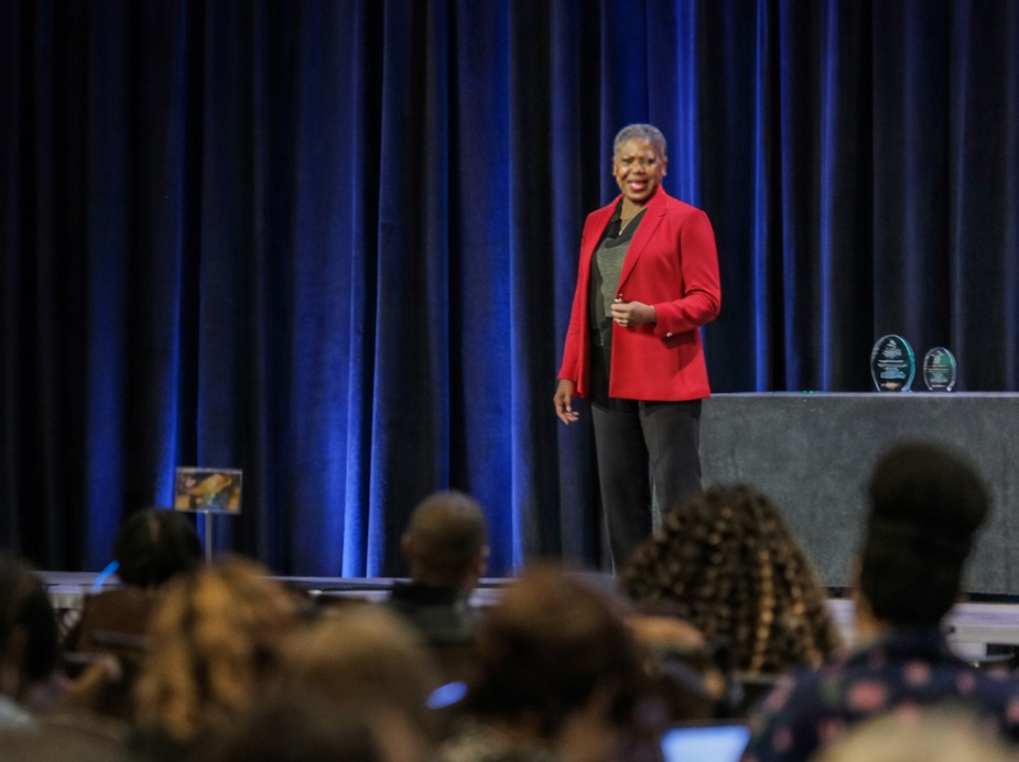 Michelle Perdue speaking on stage in a red jacket, blue curtains behind