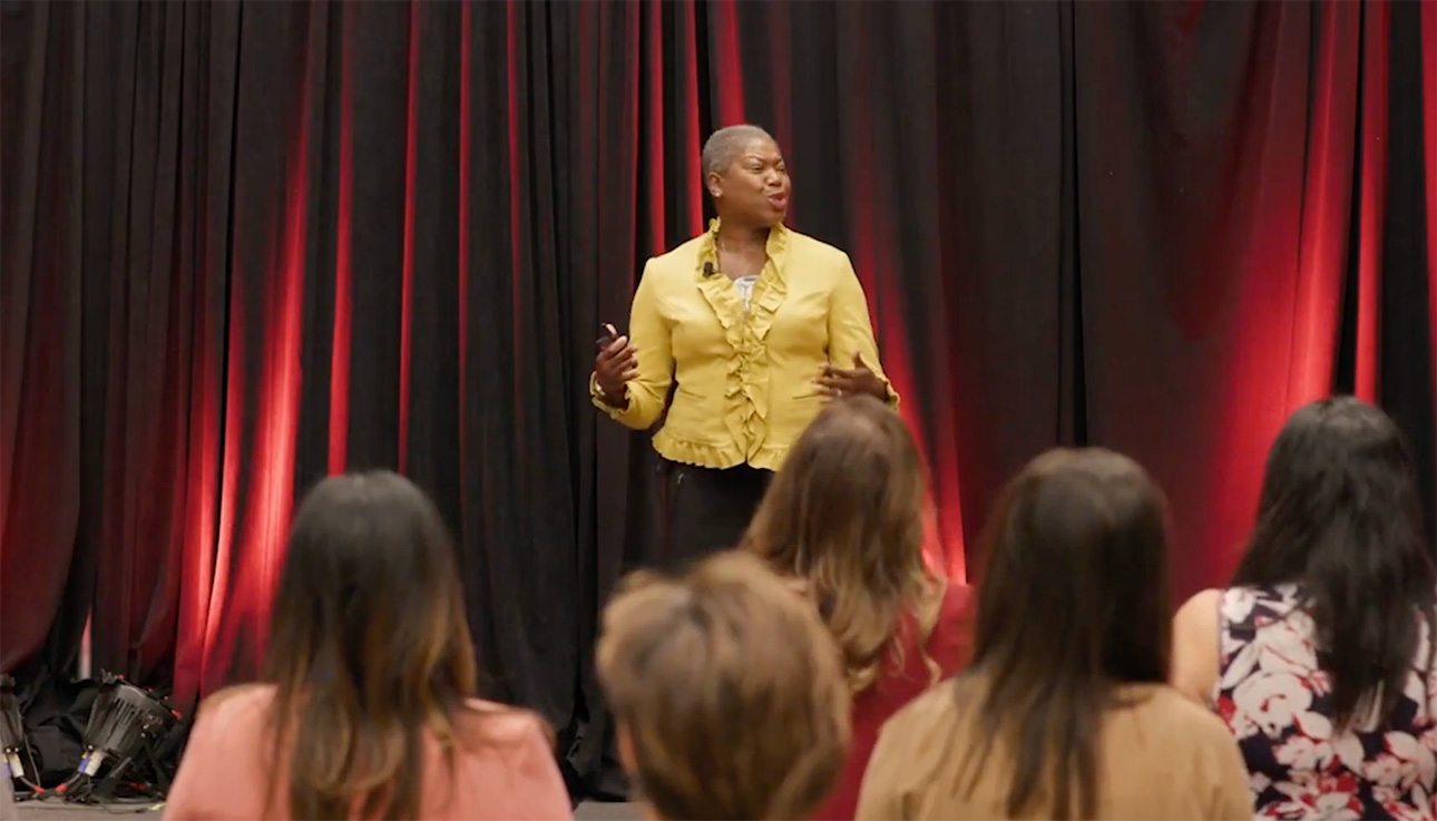 Michelle Perdue speaking and gesturing passionately in a yellow jacket with red curtains behind her on stage.